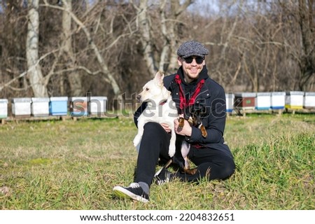 Stock photo of a smiling man sitting with his dogs in his arms in a green field, feeling peaceful on a sunny autumn day.