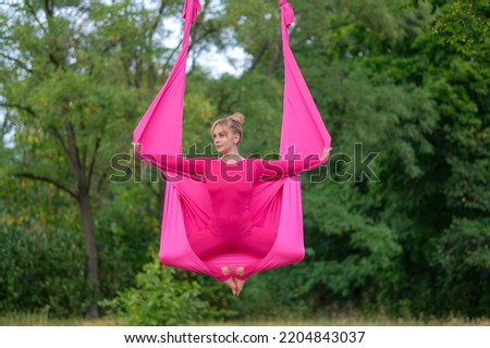 Beautiful young woman practices aerial yoga while sitting in a lotus position, in pink hammock on a tree in the park. 