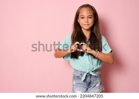 Beautiful cute little girl making heart figure with fingers standing isolated over pink background.