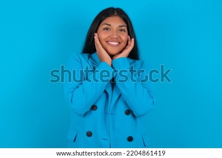 Happy Young latin woman wearing blue blazer over blue background touches both cheeks gently, has tender smile, shows white teeth, gazes positively straightly at camera,