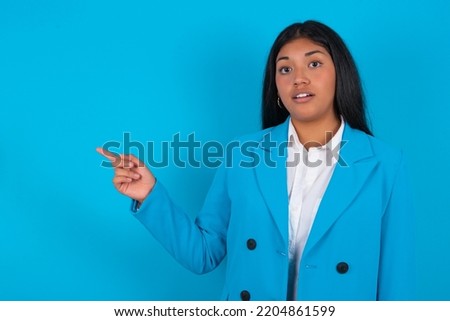 Emotive Young latin woman wearing blue blazer over blue background keeps jaw dropped from shock demonstrates amazing promo points right on blank space demonstrates big shopping sale. Advertisement
