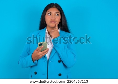 Young latin woman wearing blue blazer over blue background thinks deeply about something, uses modern mobile phone, tries to made up good message, keeps index finger near lips.