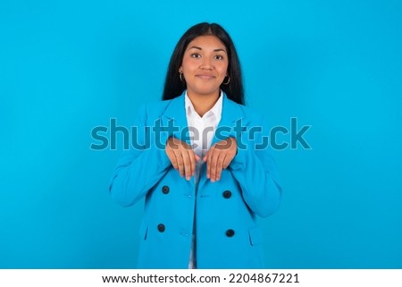 Young latin woman wearing blue blazer over blue background makes bunny paws and looks with innocent expression plays with her little kid