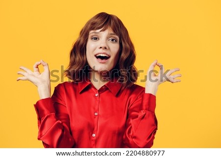 a pleasant, gentle woman stands in a stylish red shirt on a yellow background and looking into the camera happily shows the OK sign with her fingers.