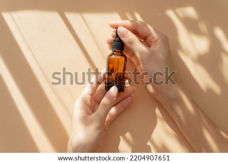 Top view of women's hands holding bottle of amber glass with cosmetic serum. Unmarked bottle for cosmetic products on beige background with floral shadow. Flat lay, copy space Royalty-Free Stock Photo #2204907651