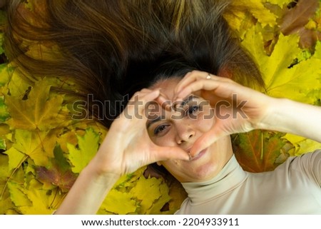 A beautiful brunette lies on yellow maple leaves and holds her hands in front of her in the form of a heart.