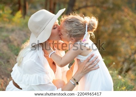 Side view of family wearing white dresses, standing on grass near trees in park forest lit up by sunset in summer. Young woman mother kissing little girl daughter with fluffy fair hair. Relationship.