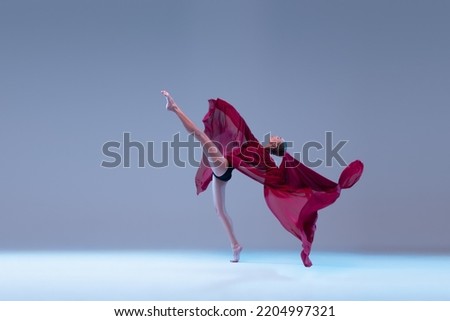 Portrait of young ballerina dancing with deep red fabric isolated over blue grey studio background. Flexibility, grace, tenderness . Concept of classic ballet, inspiration, beauty, dance, creativity
