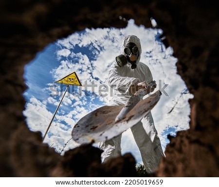 Close-up view of shovel with which scientist in gas mask digging hole to study composition of soil in danger zone. View from inside pit. On background pointing sign with human skull with crossbones.