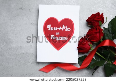 Card with love message near red roses on grey table, flat lay