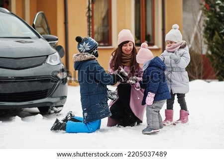 Young mother play with kids against electric car in the yard of her house at winter. Royalty-Free Stock Photo #2205037489