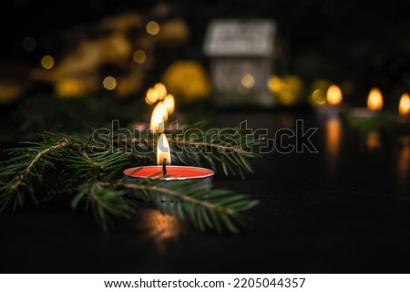 On a black table, a large candle is burning close-up, and nearby are branches of a Christmas tree strewn with small green needles. The blurred background shows Christmas lights and a toy house.