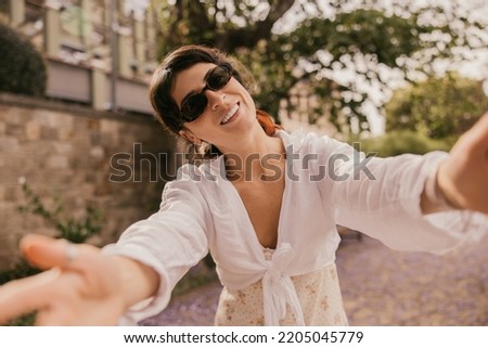 Happy young caucasian woman with snow-white smile spends time outdoors. Brunette wears black sunglasses, white summer dress. Lifestyle concept