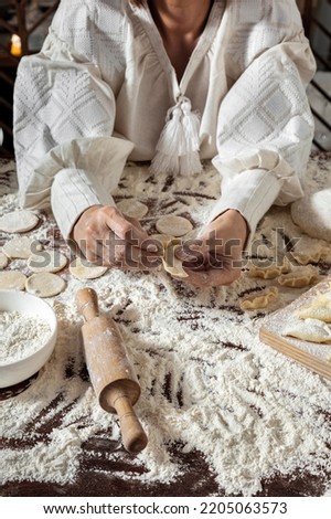 The process of making dumplings. The woman is dressed in a white, traditional Ukrainian vyshyvanka and a brown skirt. She makes dumplings on the table with her hands. Wraps mashed potatoes in round do