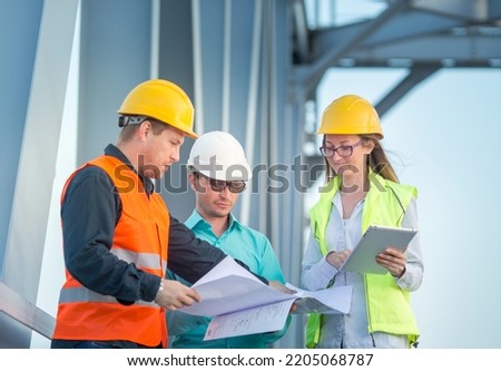 young men and a woman are standing in orange and white helmets and signal vests with documents against the background of the metal structure of the bridge. The concept of railway and road engineers.