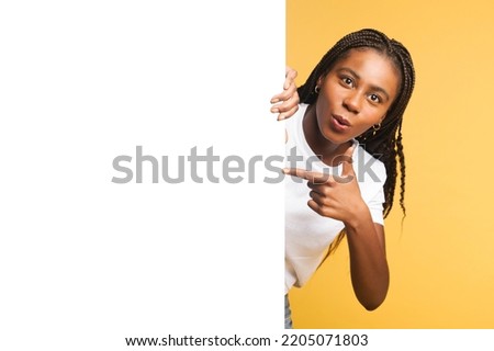 Look here. Surprised african-american woman peeps out from behind of clear empty white board and points finger on it, presenting newelty, paying your attention at blank billboard isolated on yellow Royalty-Free Stock Photo #2205071803