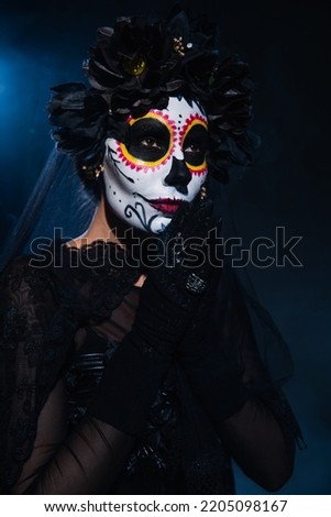 woman with halloween sugar skull makeup and praying hands looking away on dark blue background