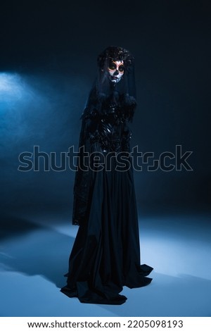 full length of woman in black witch dress and creepy halloween makeup on dark background with blue light, banner