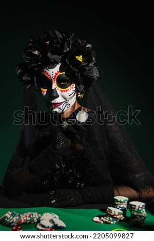 Woman in santa muerte halloween costume holding playing chip on dark green background