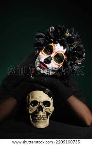 Woman in black wreath and day of death costume looking at camera near skull on dark green background