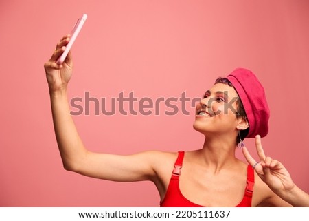 A young woman blogger with colored pink hair and a short haircut takes a picture of herself on the phone and broadcasts a smile in stylish clothes and a hat on a pink background monochrome style