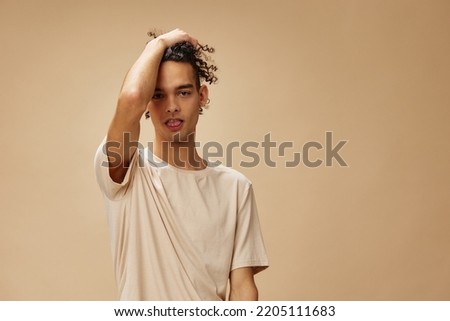 Cheerful cute awesome tanned curly man in basic t-shirt posing isolated on over beige pastel background. Fashion New Collection offer. People and Emotions concept. Free place for ad