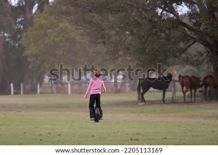 blonde girl on unicycle on polo field with horses in background Royalty-Free Stock Photo #2205113169