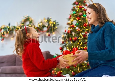 loving mother dressed in deer antlers gives gift to her daughter, at home against background Christmas tree. little girl with bow on her head is laughing happily, holding Christmas gift in her hands
