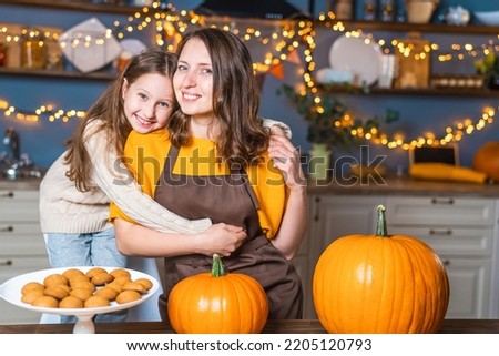 Happy Halloween. cute little girl and her mom are sitting at a table and gently hugging, looking into the frame, next to bright beautiful pumpkins. Preparation for the holiday. Maternal love and care.