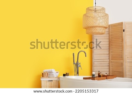 Stylish bathtub, folding screen, lamp and basket with towels near color wall