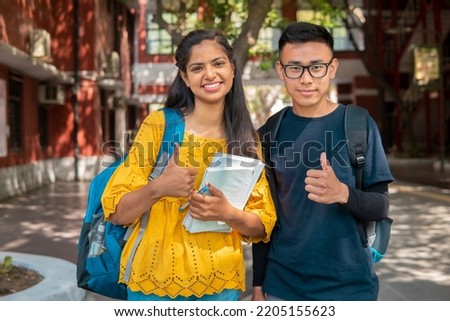 Two happy male and female college student from different ethnicity holding books in hand and showing thumbs up to the camera with smile in college campus.  Royalty-Free Stock Photo #2205155623