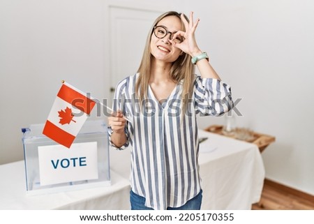 Asian young woman at political campaign election holding canada flag smiling happy doing ok sign with hand on eye looking through fingers 