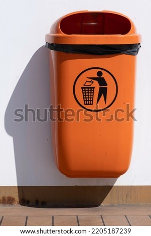 Orange plastic trash can with icon of a man throwing garbage into a basket hangs on a white wall. Drop shadow