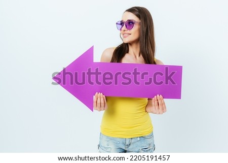 Happy young woman wearing summer top and bright sunglasses holds arrow pointing to the side and looks in same direction, standing on white studio background