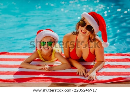 Beautiful young mother in red bikini and Christmas cap posing with cheerful daughter near the swimming pool. New Year celebration concept. Focus is at the woman. Royalty-Free Stock Photo #2205246917