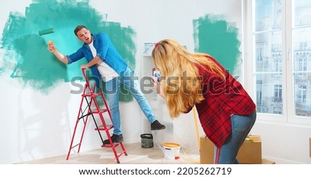 Rear of Caucasian woman standing in room taking photos on smartphone of her young handsome husband posing and painting wall in green color in good mood. Home repair, renovation and makeover concept