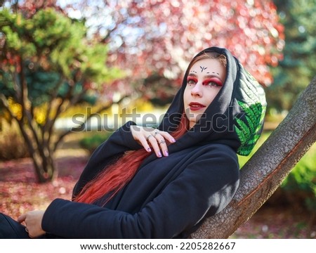 Mystical portrait of stylish red-haired girl in black coat on the background of colorful trees in the autumn garden