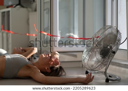 Young woman lying on floor in front of working electric fan in sunny hot summer day, enjoying cooling wind, fresh air and coolness. Girl cooling herself during hot weather at home, suffering from heat Royalty-Free Stock Photo #2205289623