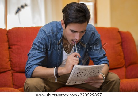Young man sitting doing a crossword puzzle looking thoughtfully at a magazine with his pencil to his mouth as he tries to think of the answer to the clue Royalty-Free Stock Photo #220529857