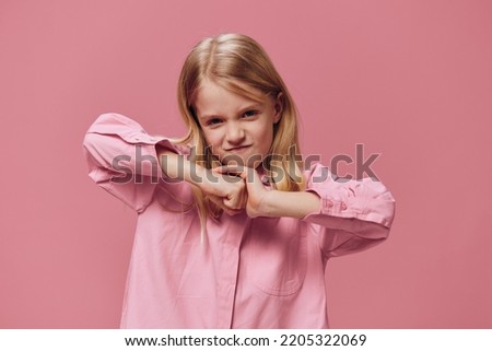 a serious, angry girl stands on a pink background in a pink shirt and holds her hands folded in fists