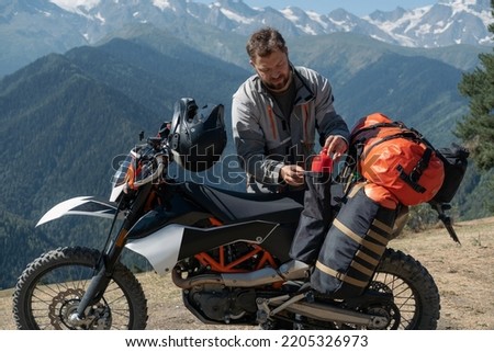 Man motorcyclist traveler standing next to dirt motorcycle and packing hiking bags with amazing mountains landscape on background 