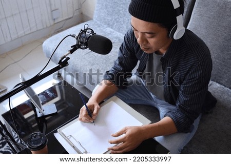 Asian man radio program host or blogger in headphones recording audio podcast using professional equipment, making notes in paper documents