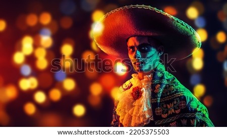 Handsome man in Mexican sombrero and traditional costume with skull makeup on his face. Festive background with colorful lights and Copy Space. Dia de los muertos. Day of The Dead. Halloween. Royalty-Free Stock Photo #2205375503