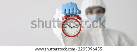Doctor in protective suit holds red alarm clock. Dangerous time spread of bacteria and viruses concept Royalty-Free Stock Photo #2205396455