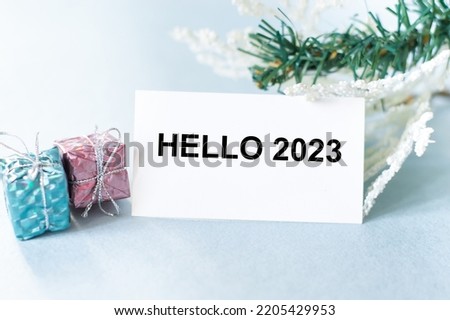Hello 2023 inscription on a white card next to spruce branches and two gifts.