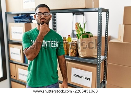 Young indian man volunteer holding donations box looking confident at the camera with smile with crossed arms and hand raised on chin. thinking positive. 