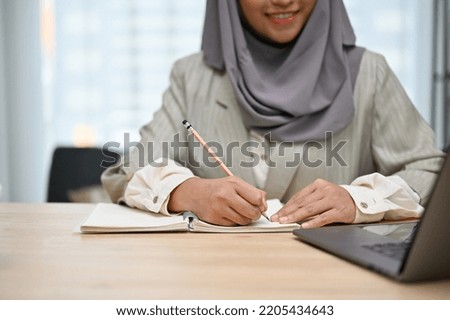 Beautiful Asian Muslim businesswoman or business employee writing her work plan on a book at her desk. cropped shot