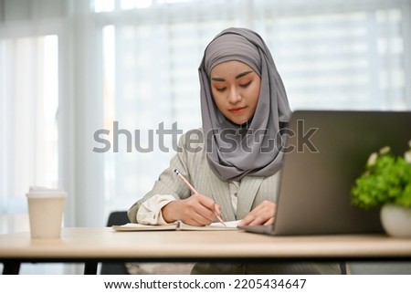 Professional and beautiful Asian Muslim businesswoman or business employee concentrating on her project assignment, writing her plan on a book at her desk.