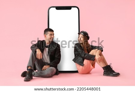 Stylish young couple sitting near big smartphone on pink background