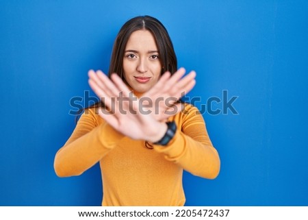 Young brunette woman standing over blue background rejection expression crossing arms and palms doing negative sign, angry face 
